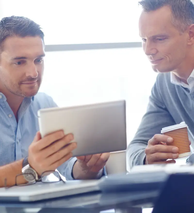 Two men in a meeting, one holding a tablet and the other a coffee cup, engaged in discussion. Light-filled modern office setting Two men in a meeting, one holding a tablet and the other a coffee cup, engaged in discussion. Light-filled modern office setting